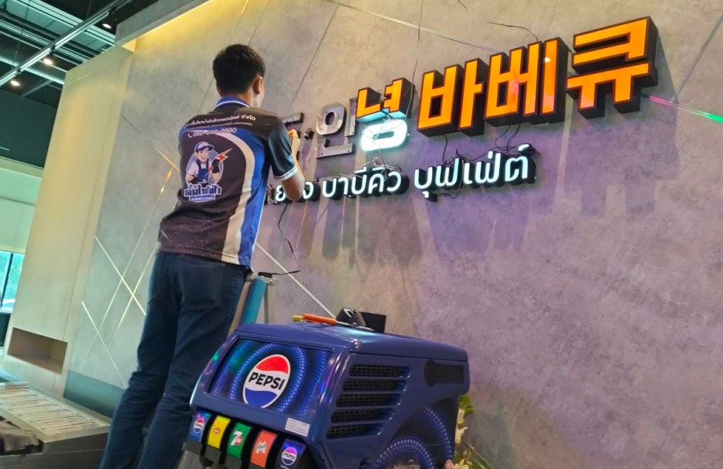 ผู้ชายกำลังติดตั้งป้ายไฟที่มีข้อความภาษาเกาหลีและไทย