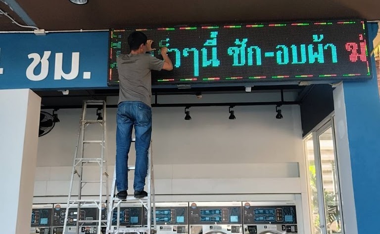 ชายคนหนึ่งขึ้นบันไดเพื่อแก้ไขป้ายโฆษณาไฟ LED ที่ติดอยู่บนผนัง