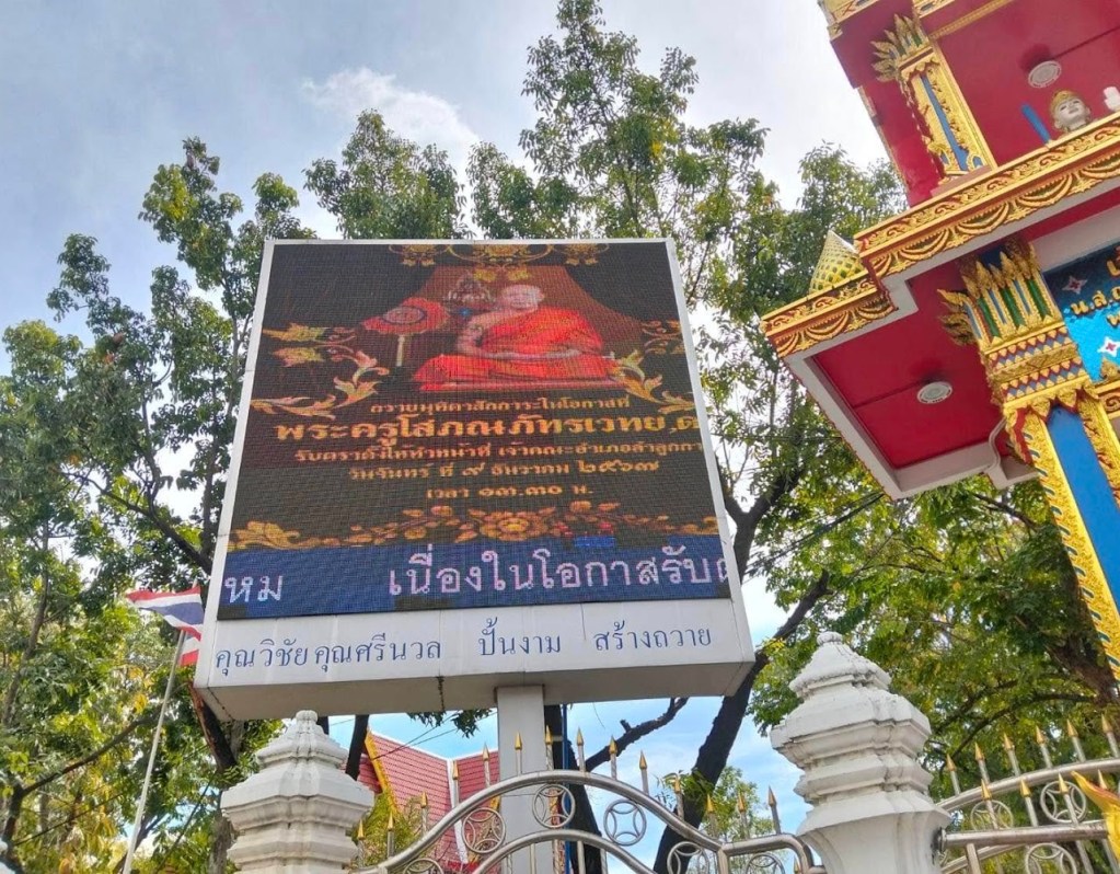 ป้ายโฆษณาแสดงภาพพระคุรุโสภณภูมิและข้อความเกี่ยวกับกิจกรรมที่เกี่ยวข้องกับการสร้างวัด ตั้งอยู่ในบริเวณที่มีต้นไม้และสถาปัตยกรรมไทย