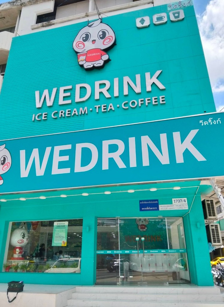 ร้าน WEDRINK มีป้ายใหญ่เป็นสีฟ้าพร้อมโลโก้ตัวการ์ตูน ไอศกรีม ชา และกาแฟ