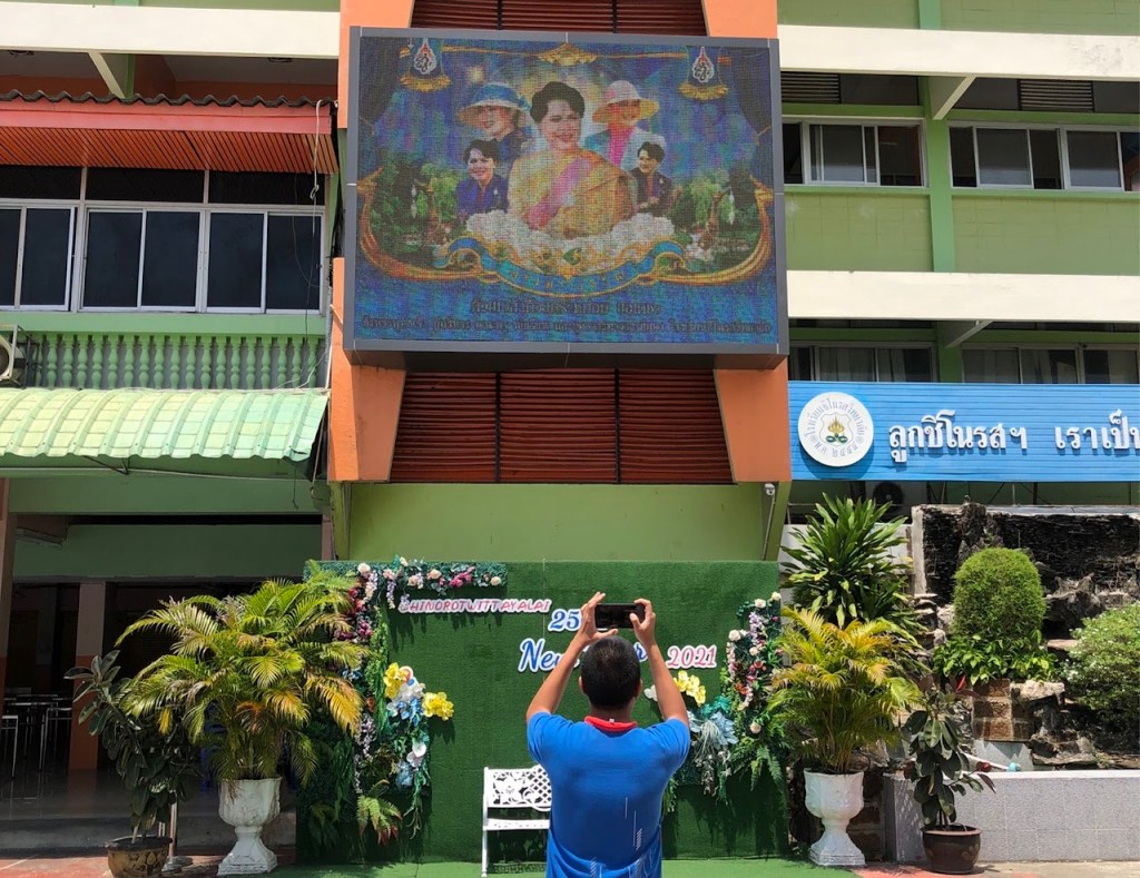 ชายคนหนึ่งถ่ายภาพหน้าจอโฆษณาที่แสดงภาพของบุคคลบนอาคาร มีการตกแต่งด้วยพืชสีเขียวและข้อความเฉลิมฉลอง